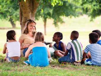 Lerares zit tegen een boom en geeft les aan kinderen.  Lerares zit tegen een boom en geeft les aan kinderen.