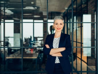 Business Kleidung – Frau im seriösen Business-Outfit steht in einem hellen Büroraum. Business Kleidung – Frau im seriösen Business-Outfit steht in einem hellen Büroraum.