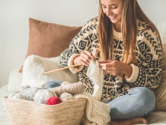 Stricken lernen – Eine Frau sitzt auf dem Bett und strickt einen weißen Schal. Stricken lernen – Eine Frau sitzt auf dem Bett und strickt einen weißen Schal.