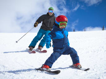 Packliste Skiurlaub – Kleiner Junge und Vater in Skiausrüstung fahren Piste hinunter. Packliste Skiurlaub – Kleiner Junge und Vater in Skiausrüstung fahren Piste hinunter.